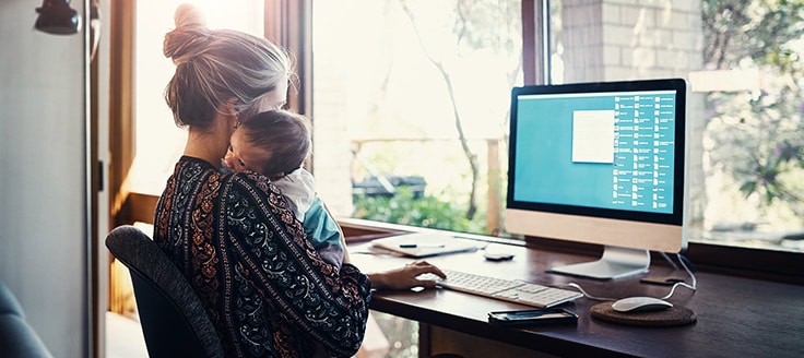 A woman works on a computer at a desk in her home office while cradling a baby on her shoulder.