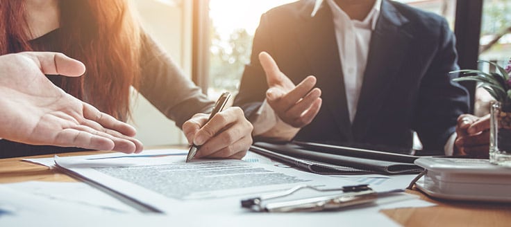 Two individuals are seated at a desk, while one signs a loan document.