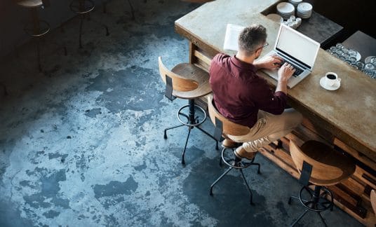 A man with glasses is seated at a desk typing on a laptop. A cup of coffee is on the man’s right.