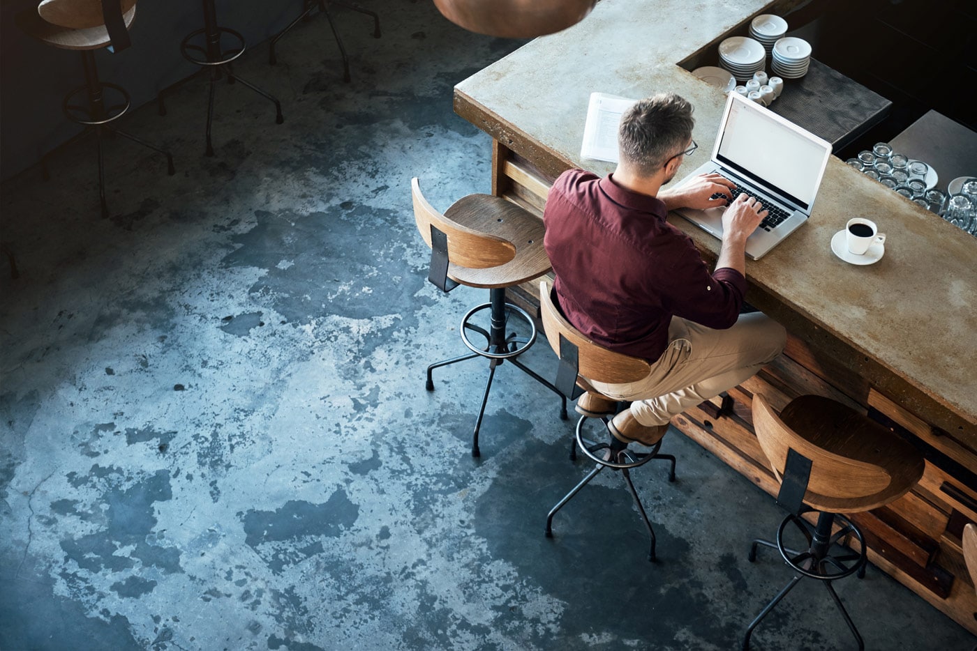 A man with glasses is seated at a desk typing on a laptop. A cup of coffee is on the man’s right.