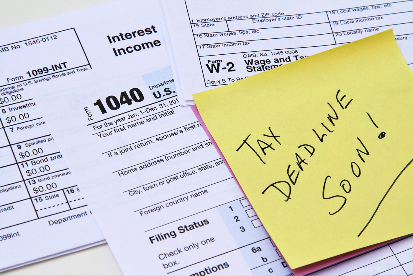 1040, W-2 and 1099-INT forms stacked with a yellow Post-It note with the words Tax Deadline Soon!