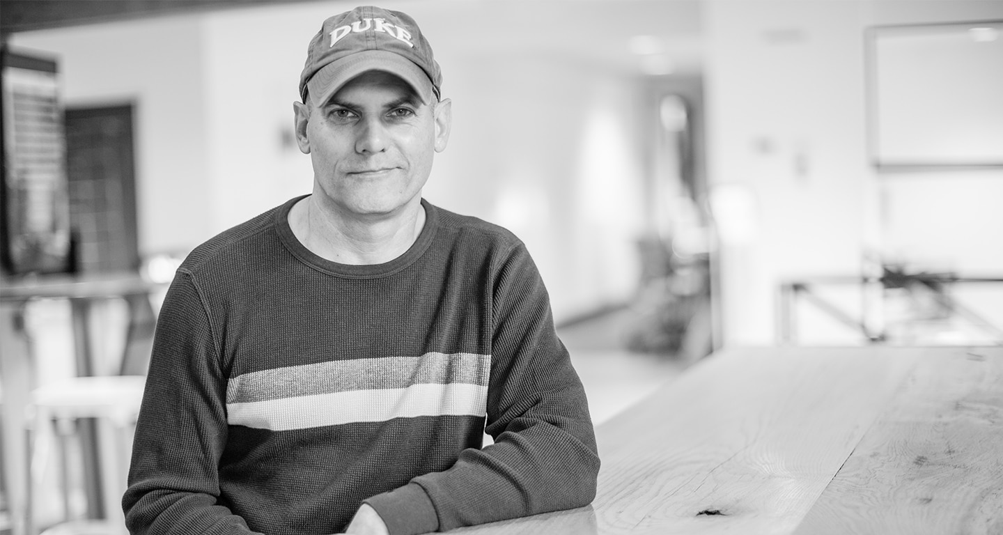 Jim Young seated at a table, wearing a duke hat and a sweater with stripes.