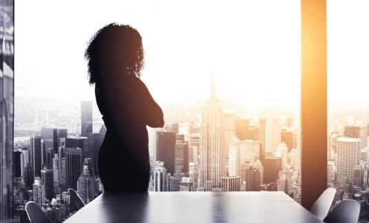 Woman in high-rise office overlooking the city out a large window.