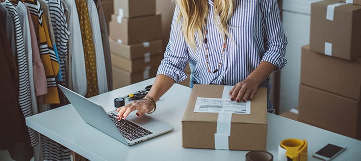 A woman consults her laptop and packages her items to mail to her Etsy customer.