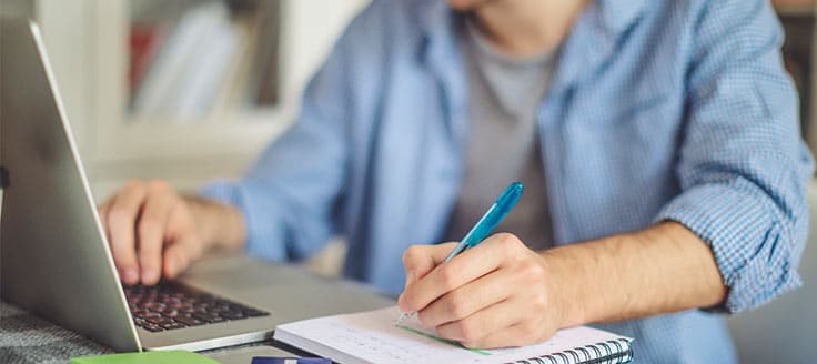 A man works at his laptop and writes in a notebook as he completes his online business course.