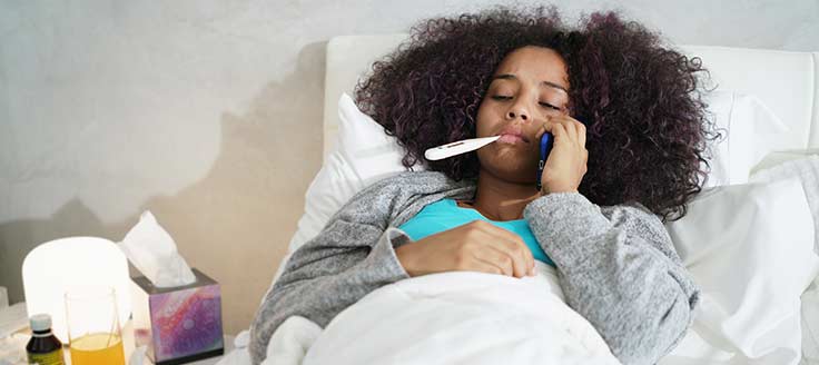A woman lies in bed with a thermometer in her mouth while she uses her cellphone to call her workplace and call out sick.