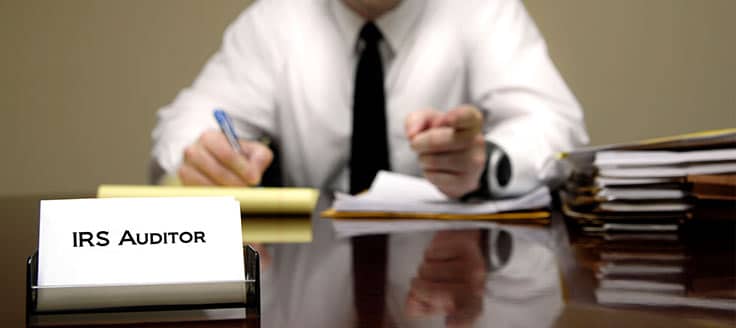 An auditor with the IRS sits at a desk and writes on a legal pad. A business card reading “IRS Auditor” sits on a stack of cards in a business card holder near the front of the desk.