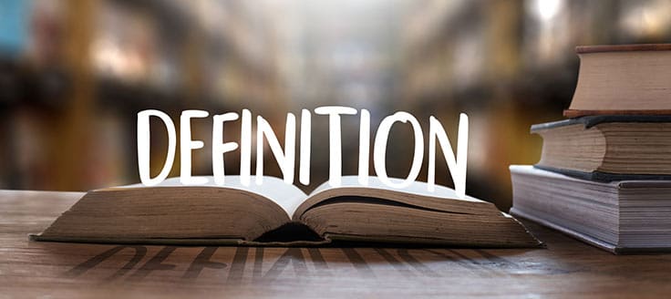 An open book sits on a table next to a stack of books. The word “definition” rests on the open book’s pages.