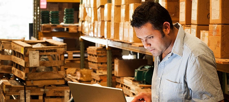 A man reviews shipping updates on his laptop as he works in a shipping warehouse.