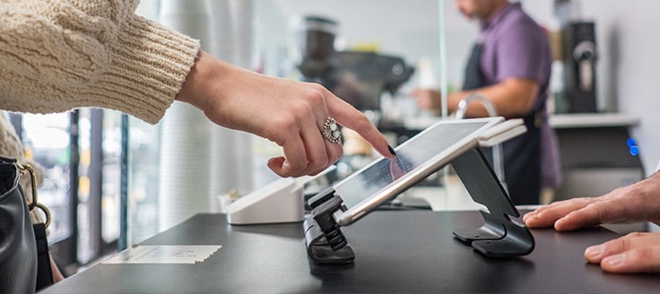 A female customer uses her finger to tap a tablet screen and complete her credit card payment processing as she purchases a coffee.