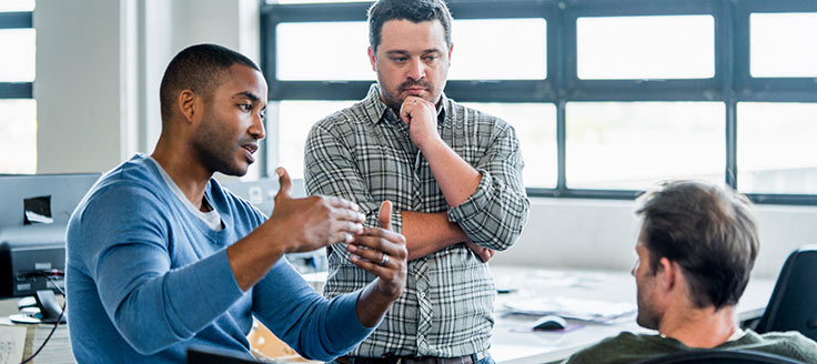 Three men have a conversation in an office. Business casual for men hinges around the tie-or-no-tie rule.