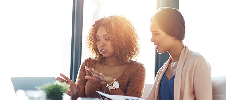 Two women review a report as they sit together at a desk in an office. Company dress codes for women typically allow for a wide range of patterns and colors.