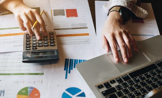 Close up of a businesswoman using a calculator and a free QuickBooks online tutorial at her desk. 