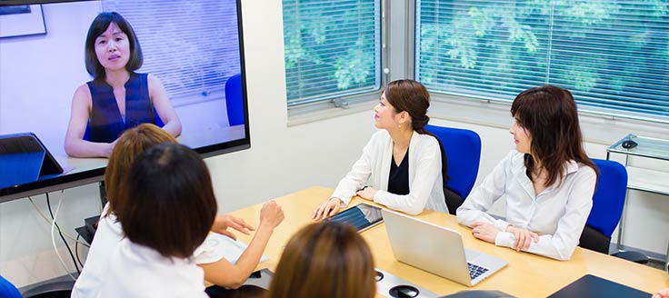 A group of co-workers sit around a table in a meeting room and watch co-worker deliver a presentation on a video screen during a teleconference.