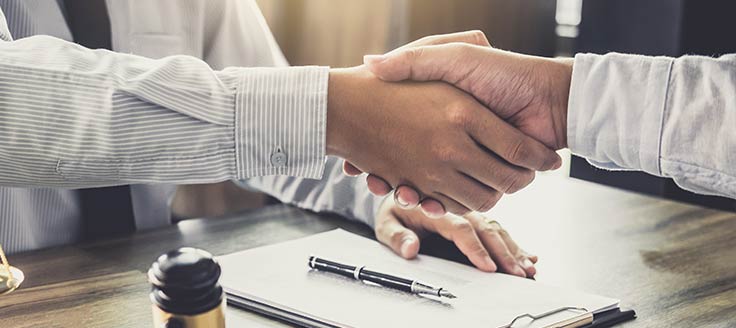 Two men in business shirts shake hands across a desk in an office. A clipboard and a pen are on the desk; it’s possibly an employment contract.