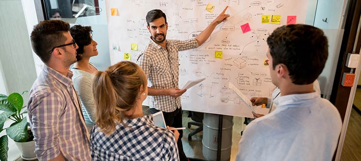 A man points to a large brainstorming board as he presents a sales plan to his co-workers during a meeting.