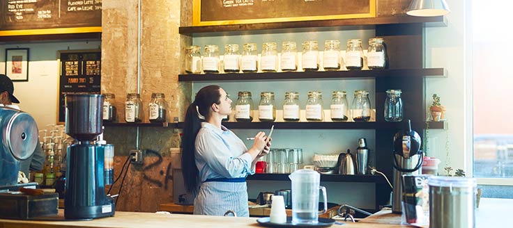 A cafe owner stands behind the counter and reviews the jars of coffee beans on shelves as she makes notes on a piece of paper. A good inventory management system helps ensure you don’t run out of stock.
