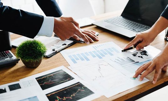 Papers cover a table’s surface in an office. A man and a woman point to different bars on an infographic report.