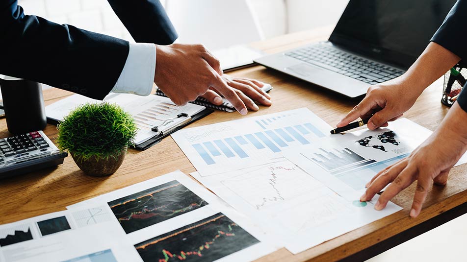 Papers cover a table’s surface in an office. A man and a woman point to different bars on an infographic report.