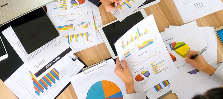 An array of reports are spread across a work table as 2 women entrepreneurs review the pages and make notes.