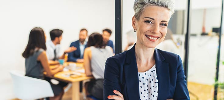 A businesswoman stands in a meeting room; her team is visible behind her.