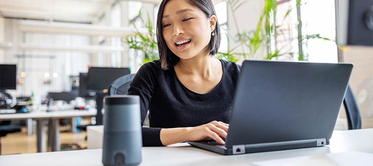 A woman at work on a laptop in an office gives a command to an AI device that's on her desk.