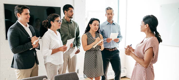 A group of new employees stand in a conference room and take notes while a female colleague speaks.