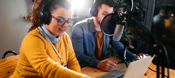 A young woman and young man record a podcast on a laptop.