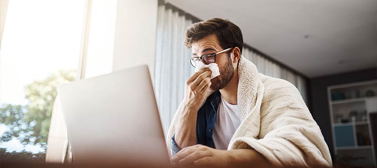 A man covered in a blanket blows his nose while working on his laptop.