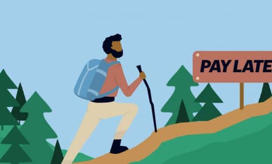 A hiker with a walking stick comes across a “Pay Later” direction sign on a trail heading up in the woods.