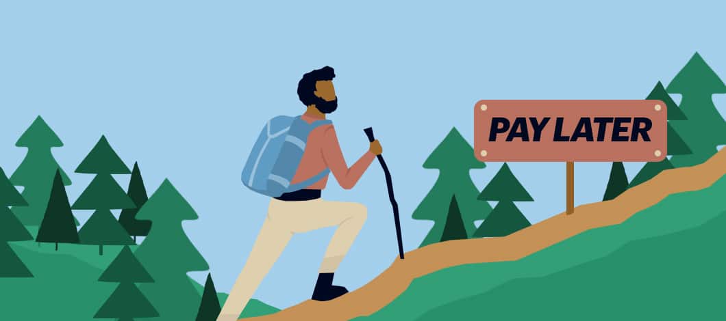 A hiker with a walking stick comes across a “Pay Later” direction sign on a trail heading up in the woods.