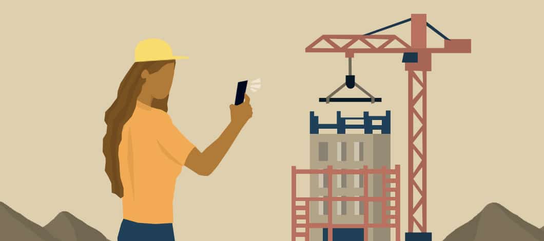 A contractor wearing a hard hat takes a photograph of a building under construction with her smartphone.