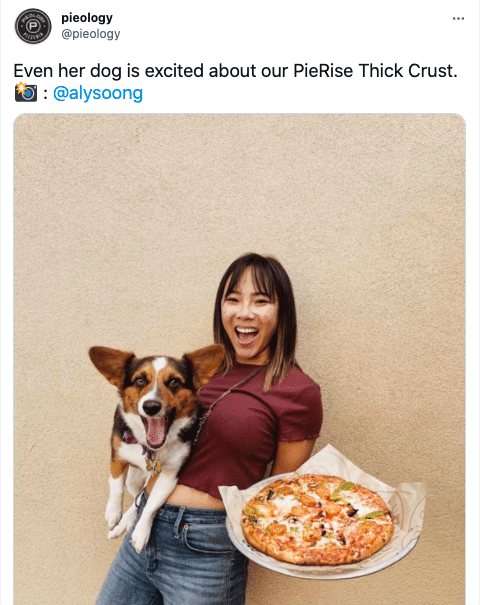 A screenshot of a restaurant's tweet featuring a photo of a woman posing with a corgi and a pizza
