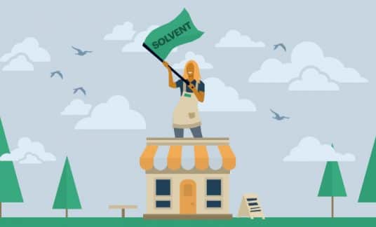 A smiling woman stands on the roof of her small shop and waving a giant green flag that reads “Solvent.”