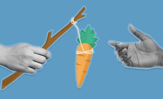 A hand reaches for a carrot tied to a stick that is being held by another hand.