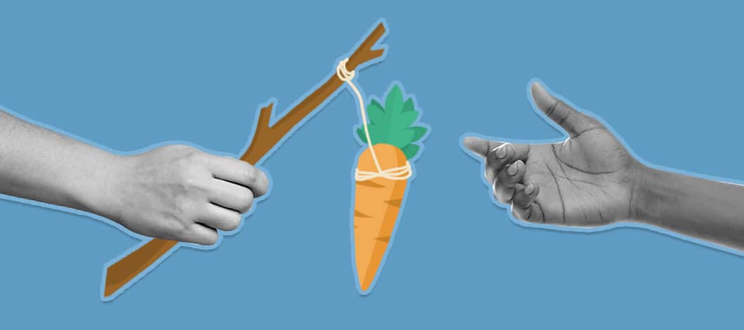 A hand reaches for a carrot tied to a stick that is being held by another hand.