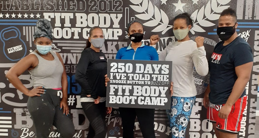 Five Masin Mills Fit Body Boot Camp clients standing for a photo opp, holding up a Fit Body Boot Camp sign