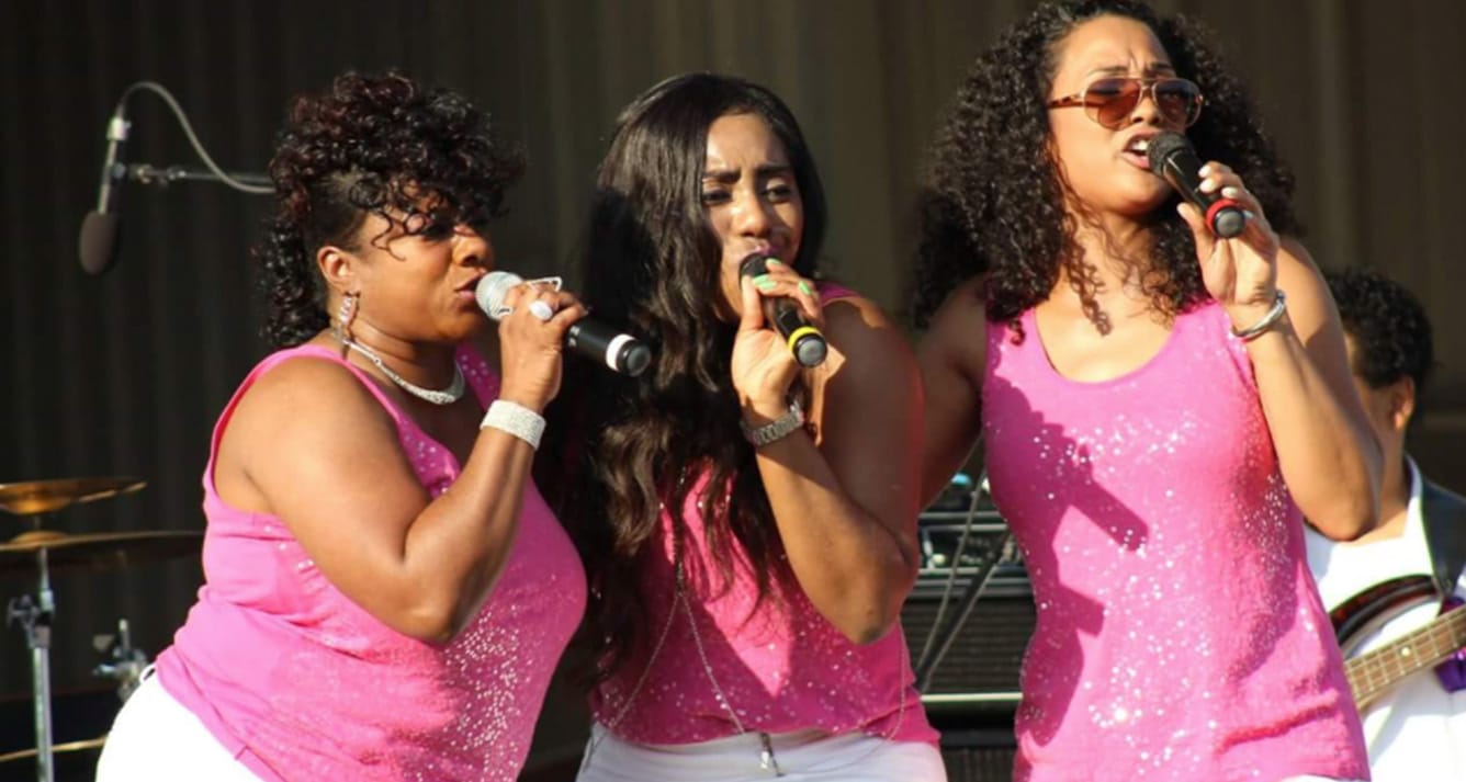 Three female singers perform with Liquid Pleasure Band onstage at a show.
